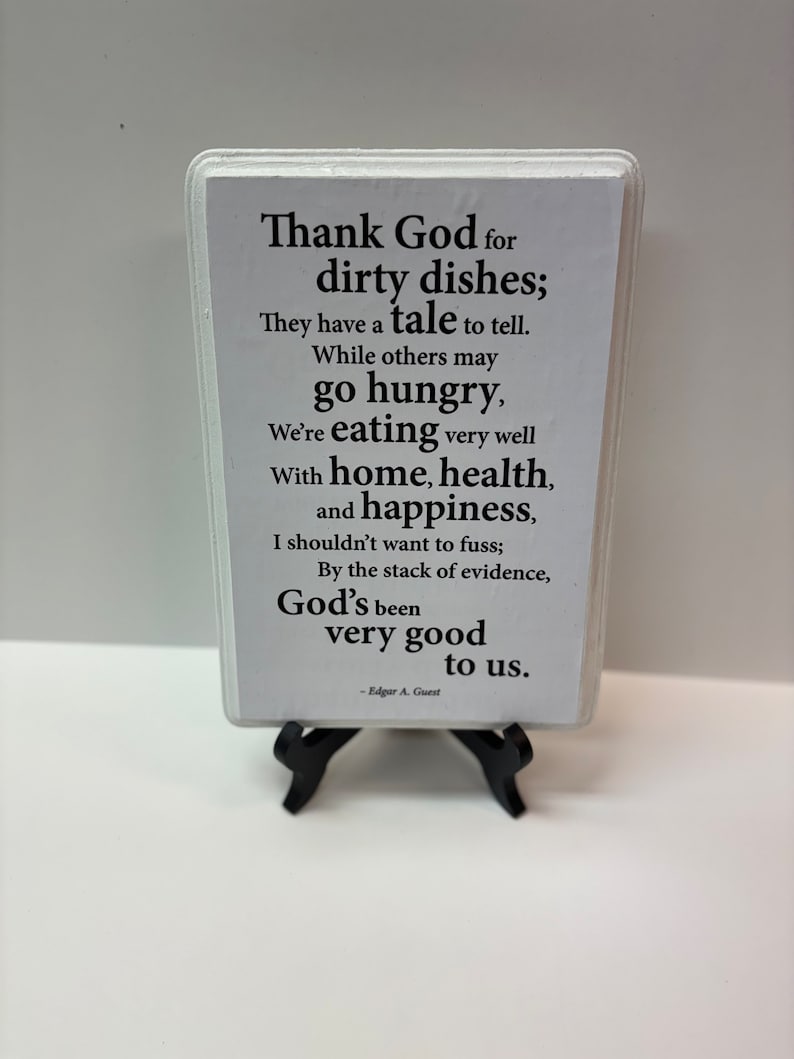May include: A white rectangular sign with black text, displayed on a black easel. The sign features a poem about gratitude for dirty dishes, home, health, and happiness, attributed to Edgar A. Guest.