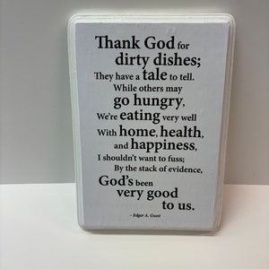 May include: A rectangular white sign with a white border, featuring black text. The text reads, "Thank God for dirty dishes; They have a tale to tell...God's been very good to us." The sign is attributed to Edgar A. Guest.