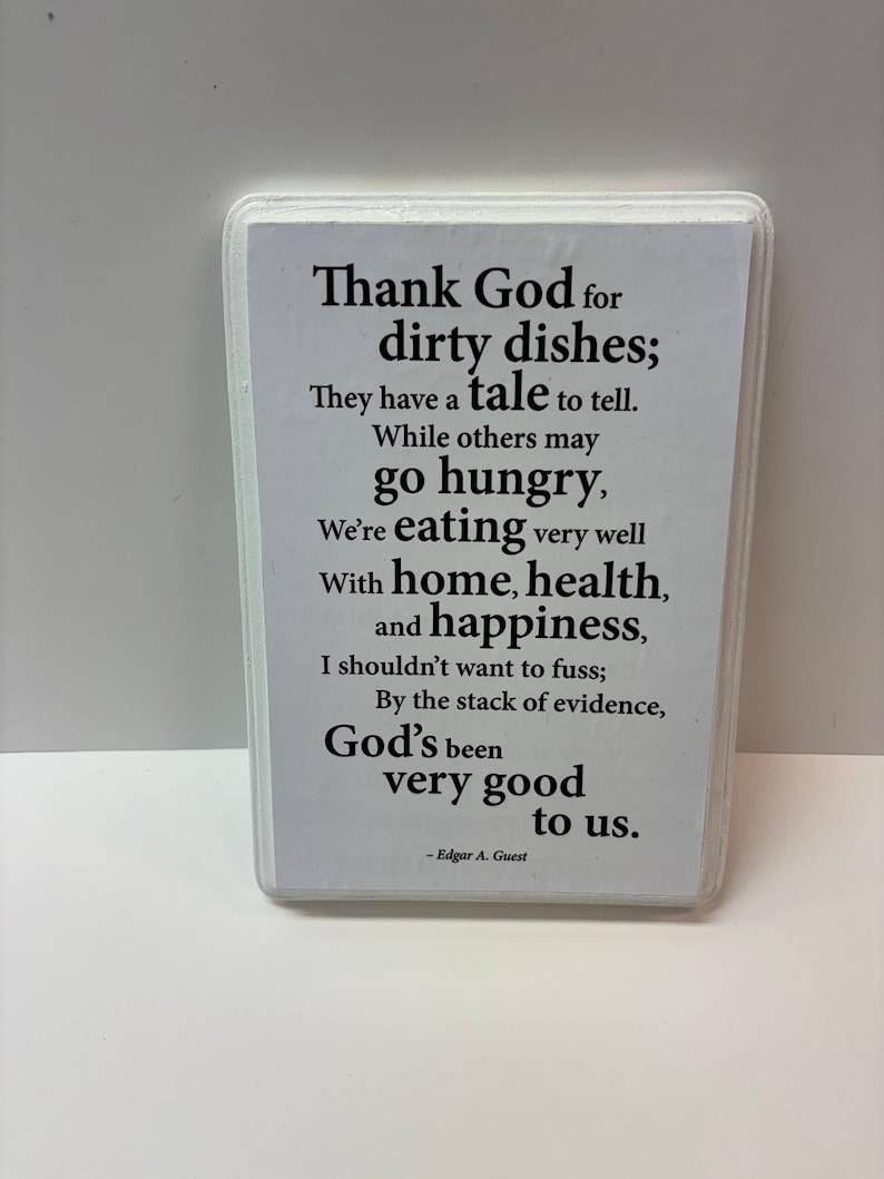 May include: A rectangular white sign with a white border, featuring black text. The text reads, "Thank God for dirty dishes; They have a tale to tell...God's been very good to us." The sign is attributed to Edgar A. Guest.