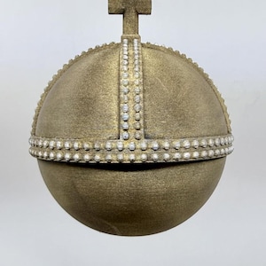May include: A gold-coloured, orb-shaped ornament with a cross on top. The ornament is decorated with a band of small, white, pearl-like beads and a vertical line of similar beads. A black cord is attached for hanging.