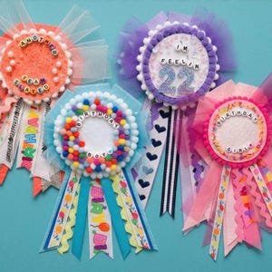 May include: Four circular birthday badges with tulle backing and hanging ribbons. One orange badge reads ANOTHER PEA SLAYED, one purple badge reads I'M FEELIN 22, one blue badge features colorful pom-poms and BIRTHDAY BOY, and one pink badge reads BIRTHDAY GIRL.