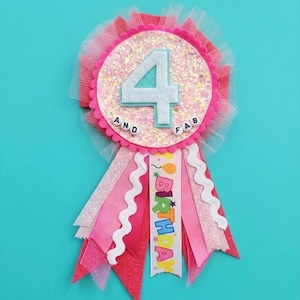 May include: A circular birthday badge featuring a glittery pink center with a large white felt number 4 and the text AND FAB. The badge is surrounded by pink tulle and has multiple hanging ribbons in pink, glittery silver, and white with the word BIRTHDAY.
