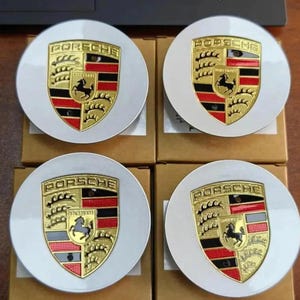 May include: Four white and gold Porsche wheel center caps. Each cap features the Porsche crest in the center, with the word "PORSCHE" above. The crest includes a black horse, red and black stripes, and gold accents.