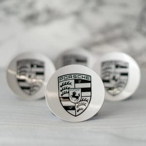 May include: A collection of silver-colored, circular objects featuring the Porsche emblem. The emblem is black and white, with the word "PORSCHE" at the top and a horse in the center. The objects are arranged on a light-colored surface.