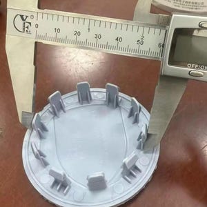 May include: A light gray automotive hub cap is measured with a digital caliper. The caliper shows a measurement of approximately 2.3 inches. The hub cap has a circular design with multiple tabs around the edge.