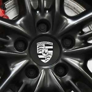 May include: Close-up of a black alloy wheel with a silver brake disc and red caliper visible. The wheel features a center cap with the Porsche logo in white on a black background. The wheel has a star-shaped spoke design.