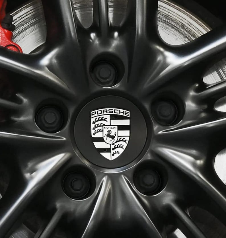 May include: Close-up of a black alloy wheel with a silver brake disc and red caliper visible. The wheel features a center cap with the Porsche logo in white on a black background. The wheel has a star-shaped spoke design.
