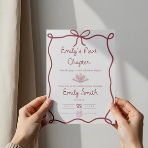 May include: A white invitation card with a burgundy border and text is held up. The text reads "Emily's Next Chapter" and invites guests to a bridal shower honouring Emily Smith on October 22nd.