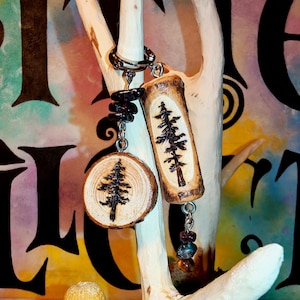 May include: Two wooden keyrings with a tree design, one round and one rectangular, suspended from a silver ring. The keyrings are attached to a light-coloured wooden branch. Black and brown stones dangle from the keyrings. The background is colourful.