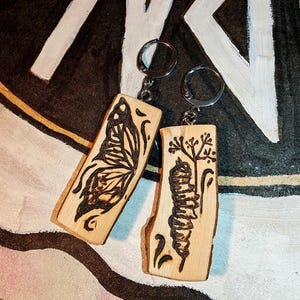 May include: Two wooden keyrings with metal hoops. One features a butterfly design, the other a caterpillar. Both are etched with dark brown lines on light brown wood.