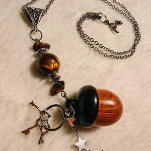 Könnte beinhalten: Halskette mit einer silbernen Kette und einem Anhänger mit einer hölzernen Eichel mit schwarzer Kappe. Die Halskette enthält auch Tigeraugenperlen, kleine Schlüssel und Sternanhänger. Die Kette ist ca. 50 cm lang.