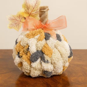 May include: A decorative pumpkin made of textured yarn in cream, yellow, and gray. It features a twine stem, an orange ribbon, and artificial fall leaves. This autumn-themed item is perfect for seasonal decor.