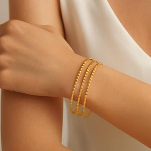 Peut inclure: Trois bracelets dorés avec un motif linéaire texturé sont portés au poignet. Les bracelets sont d'un or brillant et semblent être en métal. L'arrière-plan est de couleur neutre et douce.