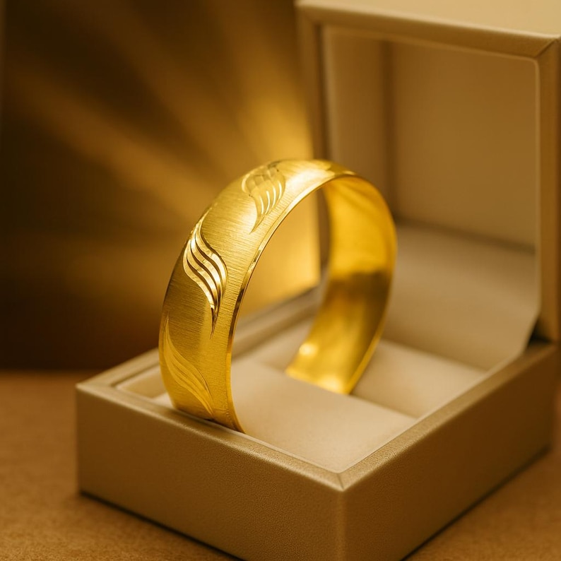 May include: A wide, gold-colored bracelet with an engraved leaf design, displayed in a beige jewelry box. The bracelet has a brushed metal texture and a bright, reflective surface. The box is open, showcasing the bracelet's design.