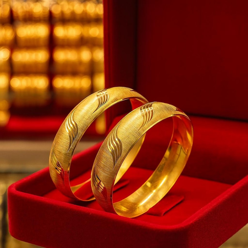 May include: Two gold bangles with a textured finish and engraved wave patterns are displayed inside a red velvet jewelry box. The bangles have a bright, shiny appearance. The background features other gold items.