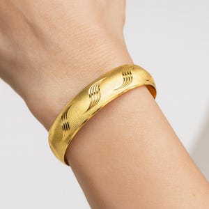 May include: A gold-colored bangle bracelet with a textured surface and etched wave-like patterns. The bracelet is worn on a wrist and has a curved, rounded shape. The jewelry is a bright, shiny gold color.