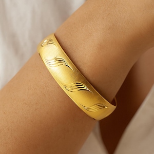 May include: A gold-colored bangle bracelet with a brushed finish and engraved wave pattern. The wide, curved bracelet is worn on an arm, showcasing its design and texture. The jewelry piece is a statement accessory.