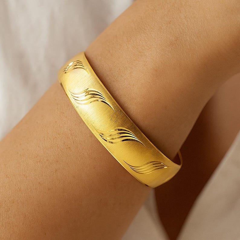May include: A gold-colored bangle bracelet with a brushed finish and engraved wave pattern. The wide, curved bracelet is worn on an arm, showcasing its design and texture. The jewelry piece is a statement accessory.