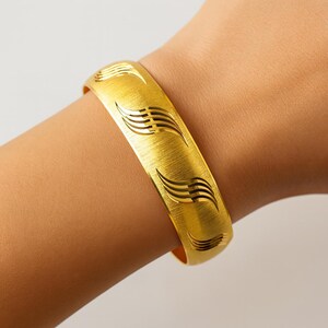 May include: A gold-colored bangle bracelet with a brushed metal finish. The bracelet features a repeating pattern of stylized wave designs. The bracelet is approximately 2.5 inches in diameter and is worn on a wrist.