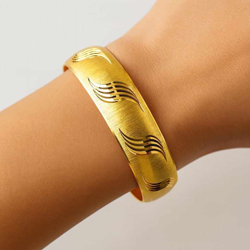 May include: A gold-colored bangle bracelet with a brushed metal finish. The bracelet features a repeating pattern of stylized wave designs. The bracelet is approximately 2.5 inches in diameter and is worn on a wrist.