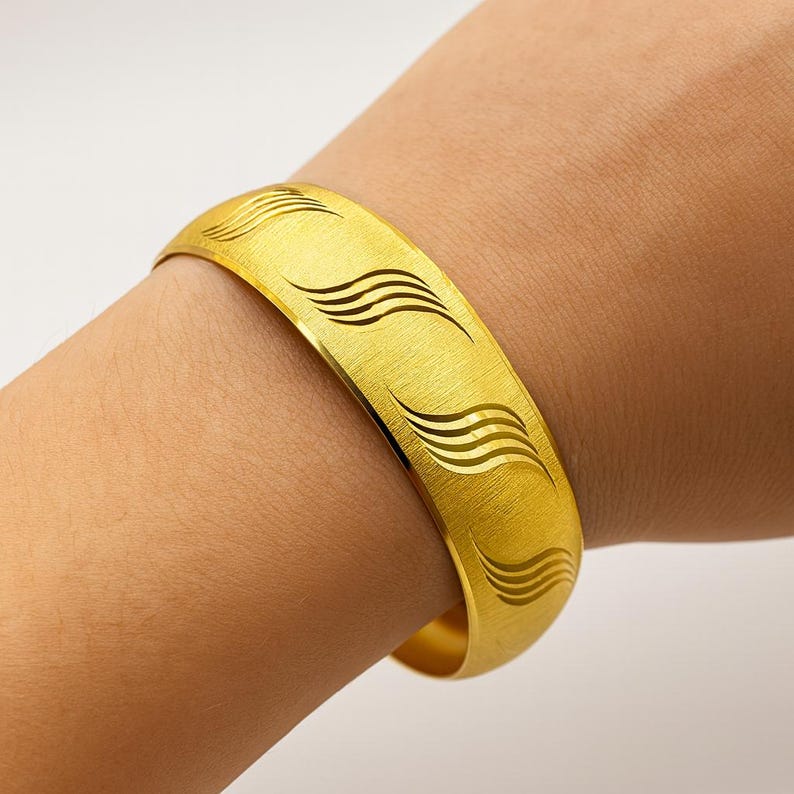 May include: A gold-colored bangle bracelet with a brushed finish and a series of wave-like designs. The bracelet is worn on a wrist and has a simple, elegant style. The bracelet is approximately 2.5 inches in diameter.