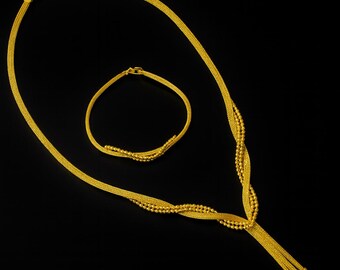 May include: A gold necklace and bracelet set against a black background. The necklace features a twisted design with beaded accents and a long, dangling strand. The bracelet has a simple, curved design with similar beaded details. Both pieces are made of gold.