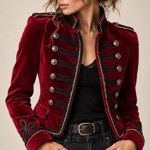 Puede incluir: Una chaqueta de terciopelo rojo intenso de estilo militar con cuello alto, hombros estructurados con charreteras y mangas largas. El frente está adornado con dos filas verticales de botones metálicos dorados, cordones negros y nudos decorativos.
