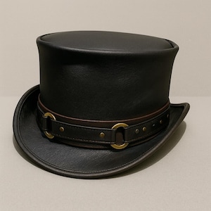 May include: A black top hat with a leather appearance. The hat features a brown band with gold-colored metal accents and rivets. The hat's brim is curved, and the crown is cylindrical.