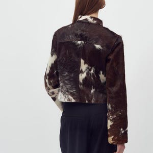 May include: A cropped jacket made of brown and white cowhide. The jacket has a collar and long sleeves. The model is wearing black pants. The jacket's pattern is a mix of dark brown and white patches.