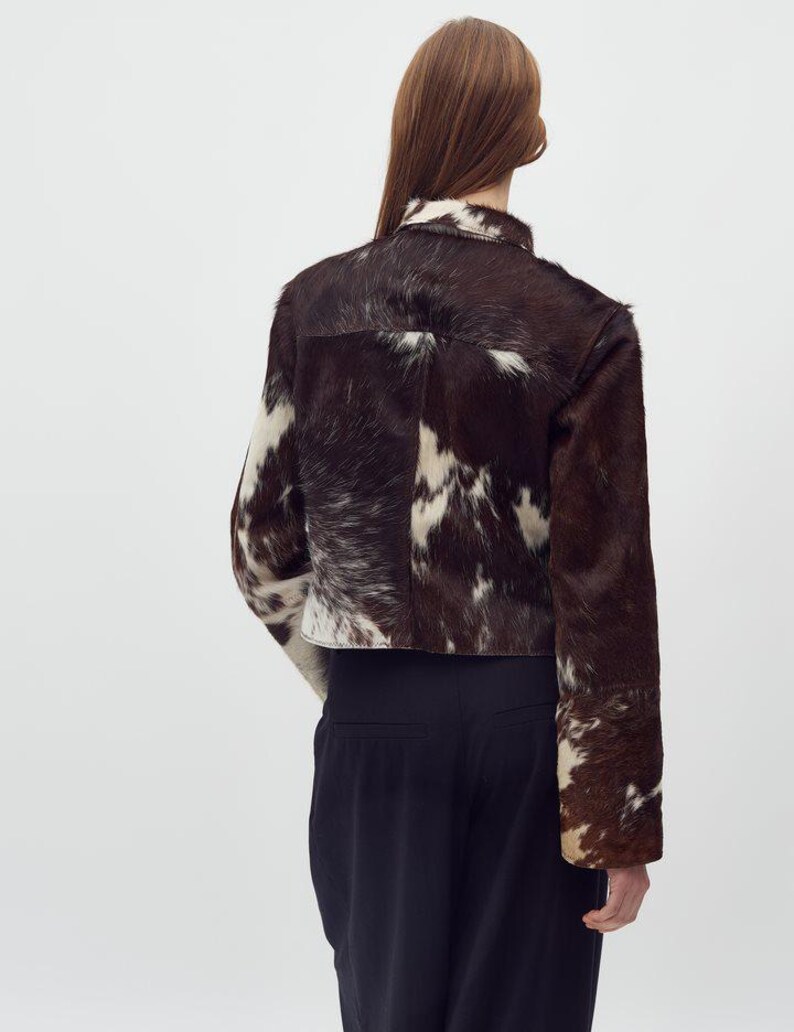 May include: A cropped jacket made of brown and white cowhide. The jacket has a collar and long sleeves. The model is wearing black pants. The jacket's pattern is a mix of dark brown and white patches.