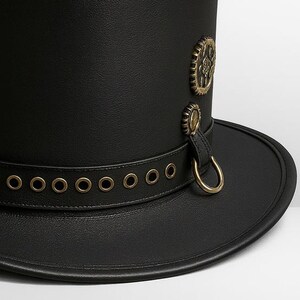 May include: A black top hat with a wide brim. The hat features a leather band with brass eyelets and a decorative brass emblem. A small leather strap with a brass ring is attached to the side.