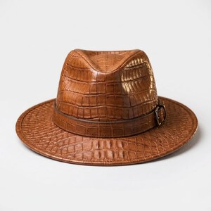 May include: A tan fedora hat featuring a textured crocodile skin pattern across the crown and wide brim. The hat includes a matching tan band around the base of the crown, secured with a small metal buckle on the side for a structured appearance.