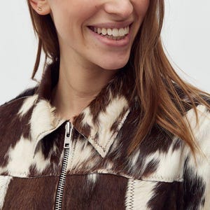 May include: A brown and white cowhide jacket with a silver zipper. The jacket has a collared neckline and is made of a patchwork design. The stitching is visible and the jacket appears to be made of a soft material.