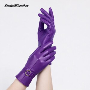 May include: A pair of vibrant purple leather gloves. The gloves have a smooth texture and a gold-colored buckle detail on the wrist. The gloves are shown against a white background. The text "StudioOfLeather" is in the top left corner.