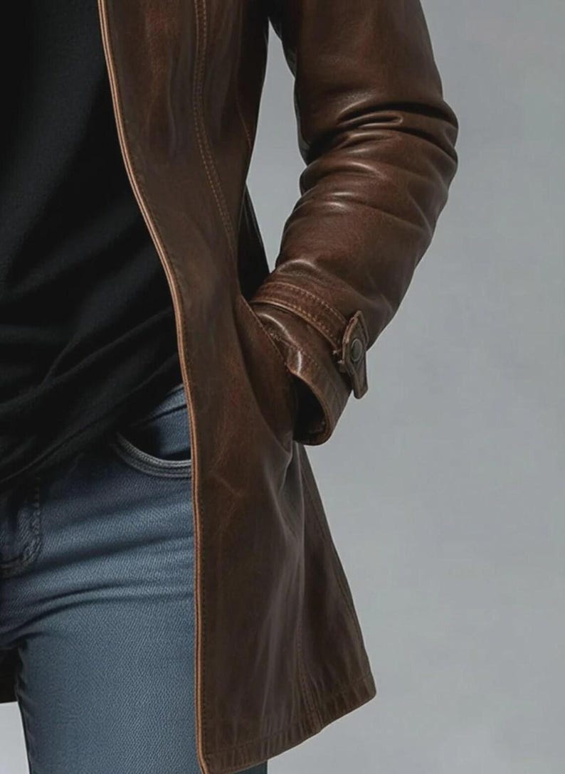 May include: A brown leather coat with a buttoned cuff detail. The coat is worn over a black shirt and blue jeans. The coat has a classic design and a long length.