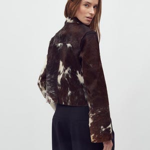 May include: A cropped jacket made of brown and white cowhide. The jacket has a high collar and long sleeves. The model is wearing black pants. The jacket's unique pattern and texture are prominent.