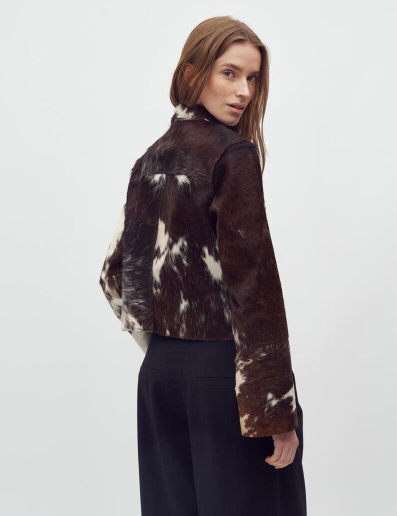 May include: A cropped jacket made of brown and white cowhide. The jacket has a high collar and long sleeves. The model is wearing black pants. The jacket's unique pattern and texture are prominent.