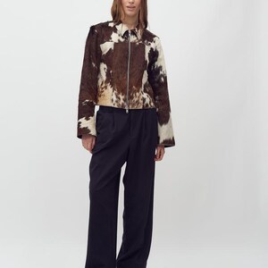 May include: A brown and white cowhide jacket with a zipper closure. The jacket is paired with dark navy blue wide-leg pants and brown leather shoes. The model is standing in front of a white background.