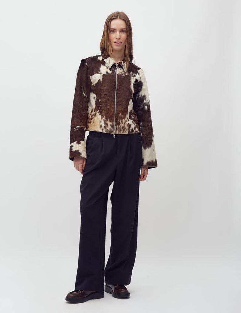 May include: A brown and white cowhide jacket with a zipper closure. The jacket is paired with dark navy blue wide-leg pants and brown leather shoes. The model is standing in front of a white background.