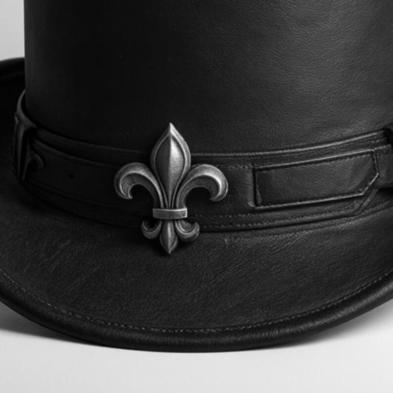 May include: A black leather top hat with a silver fleur-de-lis emblem on the band. The hat has a tall crown and a wide brim. The band is secured with a buckle. The hat is a classic accessory.