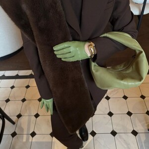 May include: A person wearing a brown suit, green gloves, and carrying a light green handbag. A brown faux fur scarf is draped over the suit. The gloves have a gold bracelet. The floor has a black and white tile pattern.