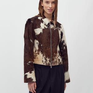 May include: A brown and white cowhide jacket with a zip-up front. The jacket has a collar and long sleeves. The model is wearing a dark blue pair of pants. The jacket is made of a soft, textured material.