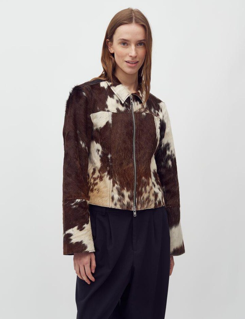 May include: A brown and white cowhide jacket with a zip-up front. The jacket has a collar and long sleeves. The model is wearing a dark blue pair of pants. The jacket is made of a soft, textured material.