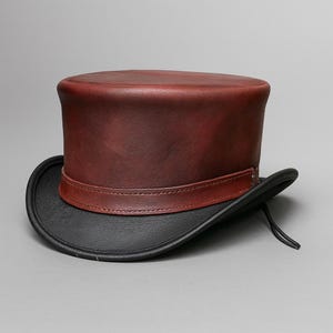 May include: A burgundy leather top hat with a black brim and a brown band. The hat has a classic, cylindrical crown and a curved brim. The hat is a formal accessory.