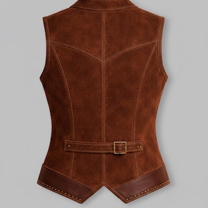 May include: A brown suede waistcoat with a notched collar and a fitted shape. The waistcoat has a belt at the waist with a square buckle and decorative stitching. The bottom hem is trimmed with darker brown leather.