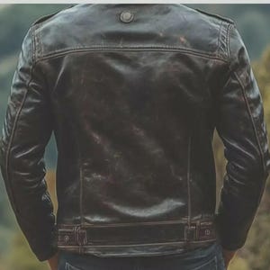May include: A dark brown leather jacket with a distressed finish. The jacket features a button detail on the shoulders, a small collar, and a belt detail at the waist. The back of the jacket is visible.