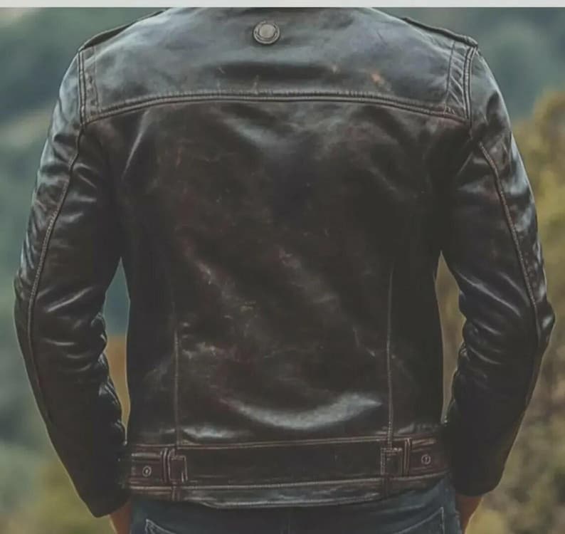 May include: A dark brown leather jacket with a distressed finish. The jacket features a button detail on the shoulders, a small collar, and a belt detail at the waist. The back of the jacket is visible.