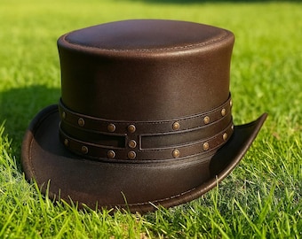 May include: A brown leather top hat with a wide brim and a decorative band around the crown. The band is adorned with small, evenly spaced rivets. The hat is set against a backdrop of green grass.