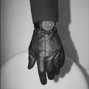 Könnte beinhalten: Schwarzer Lederhandschuh mit Armbanduhr. Die Uhr hat ein schwarzes Zifferblatt und ein schwarzes Lederarmband. Der Handschuh wird mit einem dunkelgrauen Mantelärmel getragen. Das Bild ist in Schwarzweiß.