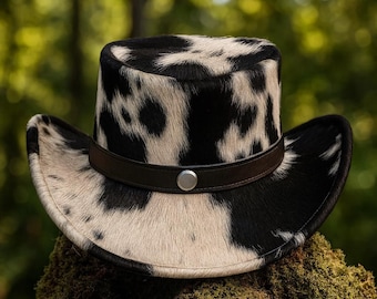 Puede incluir: Sombrero de vaquero con un patrón de manchas blancas y negras. El sombrero tiene una banda de cuero marrón oscuro con un botón plateado. El sombrero está hecho de un material texturizado y tiene un ala ancha.